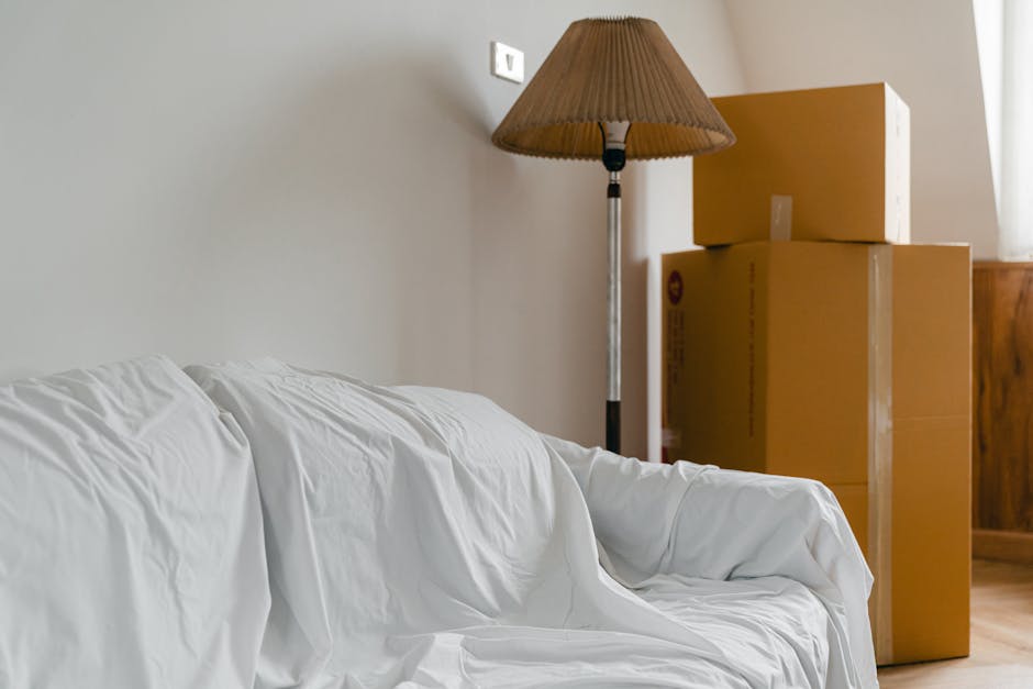 A living room scene during house relocation shows a white fabric-covered sofa positioned against a light-colored wall, with a set of three stacked cardboard moving boxes near a wooden cabinet. A tall floor lamp with a brown pleated lampshade stands beside the boxes. The boxes are sealed with packing tape and vary in size, indicating packing materials used for a move. The natural lighting comes from a nearby window, illuminating the space, while a doorway is visible on the left side of the image. The environment reflects the process of packing and preparing for furniture transport during a home relocation, with the sofa and boxes arranged for efficient loading and moving procedures. Haringey Man and Van’s services likely include handling such furniture transport and packing tasks as part of their removals process.