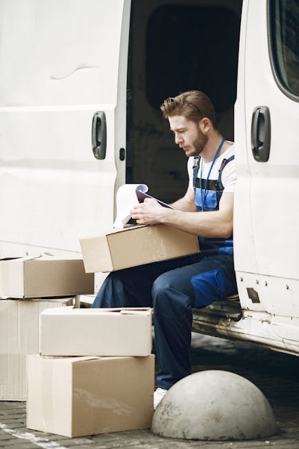 A young male worker with short brown hair and a beard, dressed in a blue and white striped t-shirt and blue overalls, seated on the edge of an open white cargo van during a home relocation. He is holding a clipboard and pen, appearing to review or record packing details. Surrounding him are several cardboard boxes of various sizes, some open and others closed, placed on the ground and on the van’s doorstep. The van is parked on a paved area, with the van door open behind him, revealing a dark interior. The scene is outdoors with natural lighting, and a concrete bollard is visible near the boxes, indicating an urban setting aligned with furniture transport and packing during a professional removal process. Haringey Man and Van is involved in house removals, including the loading process and packing logistics, as evidenced by this scene focused on organized moving activities.