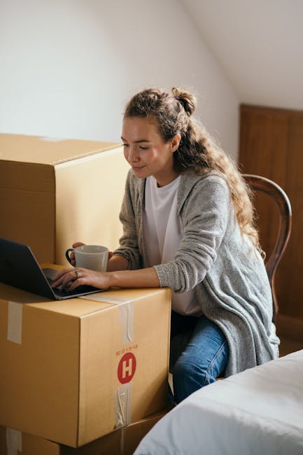 A living room scene during house relocation shows a white fabric-covered sofa positioned against a light-colored wall, with a set of three stacked cardboard moving boxes near a wooden cabinet. A tall floor lamp with a brown pleated lampshade stands beside the boxes. The boxes are sealed with packing tape and vary in size, indicating packing materials used for a move. The natural lighting comes from a nearby window, illuminating the space, while a doorway is visible on the left side of the image. The environment reflects the process of packing and preparing for furniture transport during a home relocation, with the sofa and boxes arranged for efficient loading and moving procedures. Haringey Man and Van’s services likely include handling such furniture transport and packing tasks as part of their removals process.