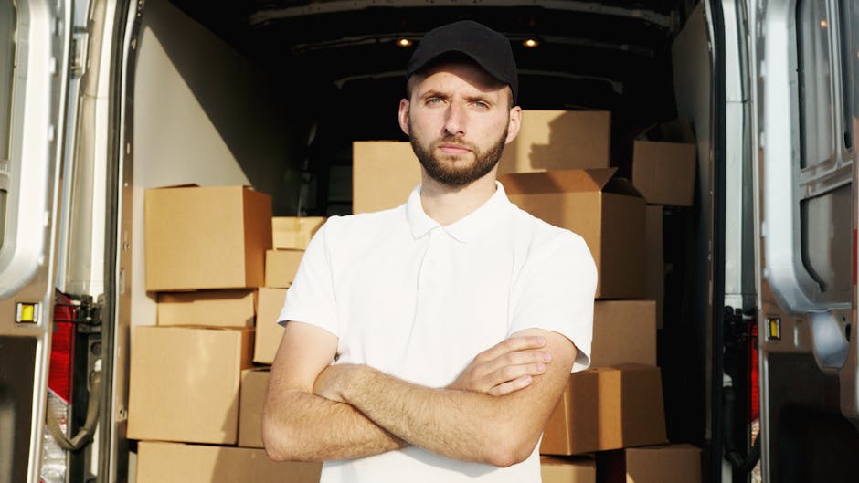 A young man with short dark hair and a beard, wearing a black cap and a white polo shirt, stands inside the rear of a moving van with the doors wide open. The van's interior is filled with neatly stacked cardboard boxes of various sizes, some wrapped in plastic. The boxes are arranged closely together, filling most of the space, with a few larger boxes positioned at the back of the van. The man has a serious expression with arms crossed as he surveys the loading process. The environment appears to be outdoors with bright natural lighting illuminating the scene. The image exemplifies home relocation and furniture transport during a packing and moving operation, illustrating the importance of proper planning to avoid mistakes that could increase costs, as highlighted in the service category of removals provided by Haringey Man and Van.