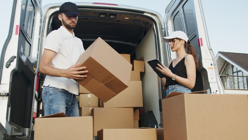 A man and a woman are standing outside the open rear doors of a white moving van parked on a residential street. The man, wearing a black cap, white t-shirt, and jeans, is holding a cardboard box while looking at the camera. Inside the van, there are several more cardboard boxes stacked neatly, along with a large piece of foam or padding material, indicating a home relocation or packing process. The woman, dressed in a white cap, black sleeveless top, and jeans, is standing beside the van with a tablet in her hands, possibly coordinating the loading or moving schedule. The loading area appears to be on a paved path with a driveway or pavement leading to the house in the background, which has a traditional design with a gabled roof and a balcony. The scene is well-lit by natural daylight, and the setup includes moving equipment such as straps or blankets, although they are not immediately visible. This image is relevant for illustrating a furniture transport or packing and moving service by Haringey Man and Van, highlighting the importance of organized loading and logistics for an efficient house removal.