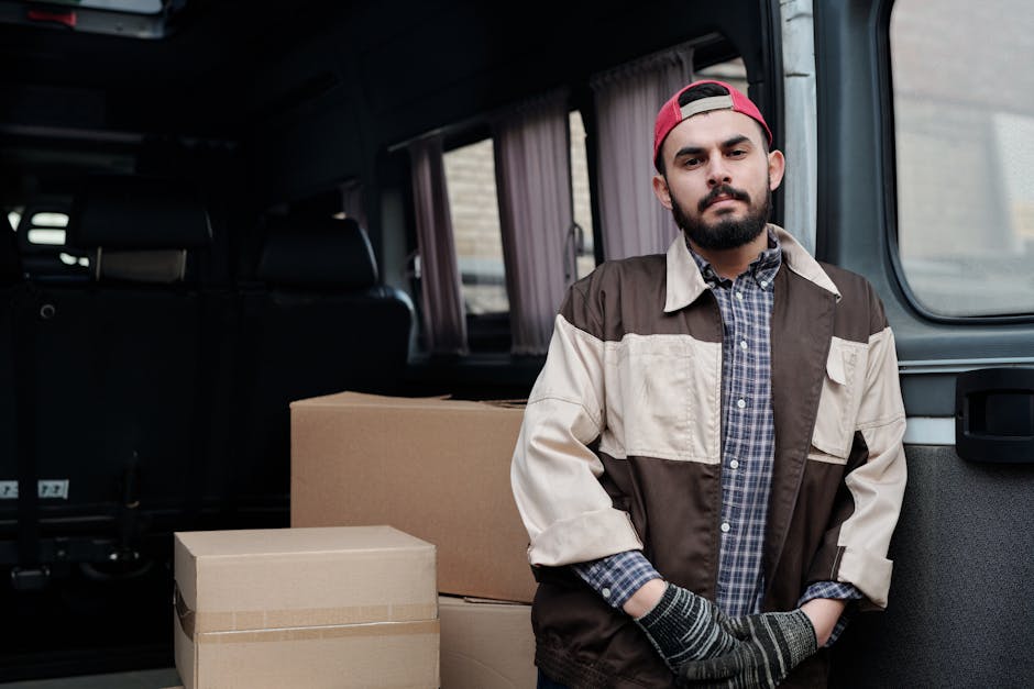 A young man with short dark hair and a beard, wearing a black cap and a white polo shirt, stands inside the rear of a moving van with the doors wide open. The van's interior is filled with neatly stacked cardboard boxes of various sizes, some wrapped in plastic. The boxes are arranged closely together, filling most of the space, with a few larger boxes positioned at the back of the van. The man has a serious expression with arms crossed as he surveys the loading process. The environment appears to be outdoors with bright natural lighting illuminating the scene. The image exemplifies home relocation and furniture transport during a packing and moving operation, illustrating the importance of proper planning to avoid mistakes that could increase costs, as highlighted in the service category of removals provided by Haringey Man and Van.