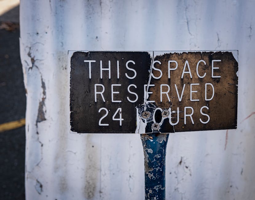 A close-up image of a weathered metal parking restriction sign mounted on a pole, with peeling paint and rust visible. The sign reads 'THIS SPACE RESERVED 24 HOURS' in white letters against a black background, though parts of the sign are damaged and partially obscured. The sign appears to be affixed to a white wall that shows signs of peeling paint and weathering. This image relates to parking regulations that may impact house removals and moving logistics, which Haringey Man and Van provides assistance with in accordance with Haringey Council rules for removals parking permits. The setting suggests an urban environment where careful planning of vehicle parking is essential for home relocation and furniture transport tasks.