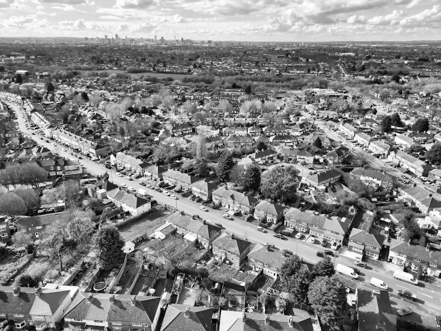 Black-and-white aerial photograph of a residential neighbourhood showing a row of terraced houses along a main street, with several delivery vans and cars parked on the road. Some houses have small front yards with fences, gardens, and trees. The street appears to be a typical UK suburban area, with rooftops, chimneys, and paved pavements visible. In the background, an expansive urban skyline with taller buildings and distant cityscape can be seen under a partly cloudy sky. This image captures the scene of home relocation activity, where vehicles are parked close to houses, suggesting loading or unloading processes involved in house removals. Haringey Man and Van provides removals services, including furniture transport and packing support, which are often conducted in areas like this during same-day relocation or moving logistics.