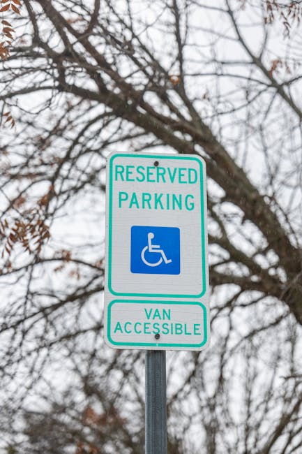 A vertical rectangular sign mounted on a metal pole outdoors, featuring a white background with green and blue text. The upper part displays 'RESERVED PARKING' in green capital letters with a blue wheelchair symbol beneath, indicating designated accessible parking. Below, the sign reads 'VAN ACCESSIBLE' in green text. The background shows a cloudy sky and leafless tree branches, suggesting a winter or late autumn setting. This signage is commonly seen in urban areas, including residential streets or parking lots, to designate accessible parking spaces and inform about van access. The context relates to parking regulations relevant to house removals or relocation services, such as those provided by Haringey Man and Van, supporting efficient planning for home moving logistics and equipment access.