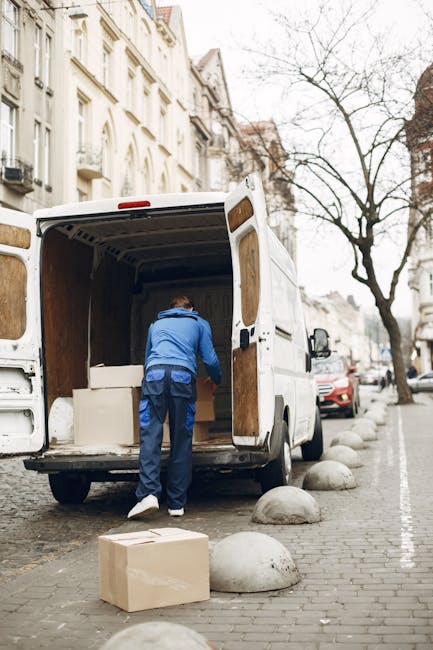 A man wearing a blue jacket and dark trousers is loading a cardboard box into the back of a white moving van parked on a city street. The van's interior shows empty wooden paneling, and the side door