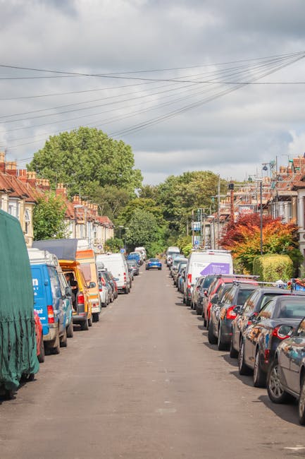 Photograph of a residential street in the Seven Sisters N15 area showing a row of parked cars on both sides of the narrow asphalt road, with some parked vehicles covered in protective fabrics for home relocation purposes. The street is lined with terraced houses featuring brick facades, bay windows, and small front gardens with shrubs and trees, including a large green-leafed tree at the end of the street. Overhead, electrical wires run across the scene, and the sky is partly cloudy with patches of sunlight. The scene depicts typical urban surroundings where careful parking, parking permits, and moving logistics are important, supporting services such as removals and furniture transport offered by Haringey Man and Van, especially when planning house moves or packing and loading processes in the area.