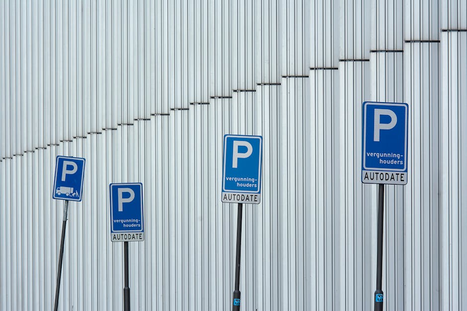 Four blue parking permission signs positioned on black metal poles in front of a large, vertically ribbed metallic wall, with one sign showing an icon of a van. The signs include the letter 'P' and the words 'vergunning-houders' and 'AUTODATE' on a white background. The signs are arranged in a slight arc, with the tallest sign at the center and shorter signs on either side. The environment appears to be an outdoor parking area or designated loading zone, possibly associated with house removals or moving services, as suggested by the context of the webpage. The lighting is even, emphasizing the metallic surface and the signs, which indicate authorization for parking or loading, relevant to relocation logistics.