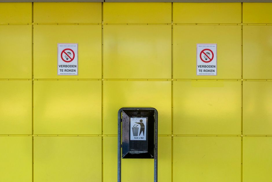 A yellow wall with a grid pattern of square panels, each with visible screws at the corners. Two signs displaying a no-smoking symbol with the text 'VERBODEN TE ROKEN' are mounted on the wall, positio