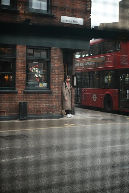 A person wearing a beige coat, dark trousers, and white sneakers standing on a wet street pavement outside a brick building with black-framed windows, amidst a rainy day. Behind them, a double-decker red bus is parked close to the curb, partially visible in the frame. The building features a sign indicating 'Lordship Road' above the windows, and a sticker on one window advertising pizza. The scene reflects urban surroundings typical of a house removal or home relocation process, with the bus possibly used for transporting furniture or belongings during a move. The street is damp, indicating recent or ongoing rain, and the environment appears quiet and functional for moving logistics, supported by the presence of a person possibly waiting or involved in a packing or loading activity associated with house removals.