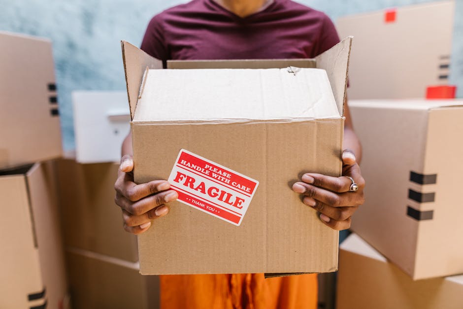 A person wearing a maroon shirt holding an open cardboard box labeled 'Fragile, Handle with Care' in red and white stickers, inside an indoor storage or moving environment. The individual’s hands are visible, with one wearing a ring on the ring finger. In the background, there are several closed cardboard boxes stacked on top of each other, some with black tape sealing the flaps, indicating an organized packing process. The setting suggests preparations for house relocation or furniture transport, with visible materials such as cardboard and packing tape. The lighting is bright and even, highlighting the focus on the cardboard box and the careful handling involved in packing and moving applications, consistent with services offered by Haringey Man and Van.