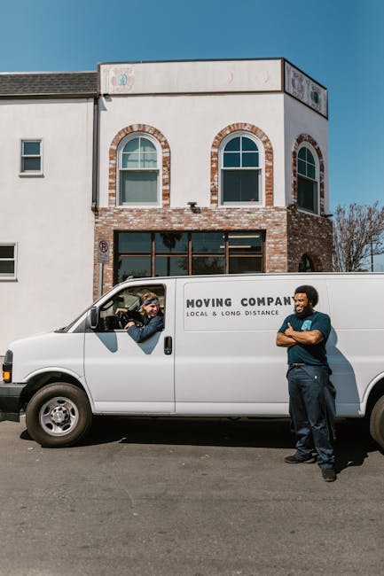 A white moving van with the branding 'MOVING COMPANY LOCAL & LONG DISTANCE' parked in front of a two-storey residential building with a white facade and brick accents around the arched windows. Inside the van’s cab, a person wearing sunglasses and a beanie is visible, engaged in conversation with another person standing outside. The person outside, dressed in a dark shirt and trousers, is leaning against the van with arms crossed, observing the loading process. The building’s ground floor features large glass doors and windows, while the upper floor has two arched windows. The scene is set outdoors on a paved street during daylight with clear weather, and the building’s exterior suggests a typical urban or suburban residential area. This image relates to house removals and furniture transport, highlighting the preparation and engagement involved in home relocation services presented by Haringey Man and Van.