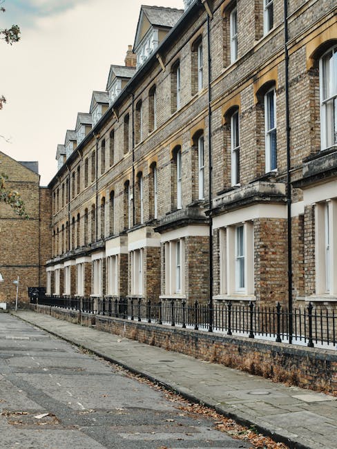 A row of traditional Victorian terraced houses constructed from yellowish-brown brick, featuring white-framed sash windows and small front gardens enclosed by black wrought iron fences. The pavement runs alongside the buildings, with some fallen leaves scattered on the ground and a narrow alleyway visible at the end of the street. The homes are situated on a slightly inclined street under an overcast sky, with no visible vehicles or people present. This residential setting reflects typical urban housing styles often encountered in neighbourhoods requiring home relocation or moving services, such as those provided by Haringey Man and Van, especially when navigating tight or narrow Victorian homes during packing, furniture transport, or loading processes.